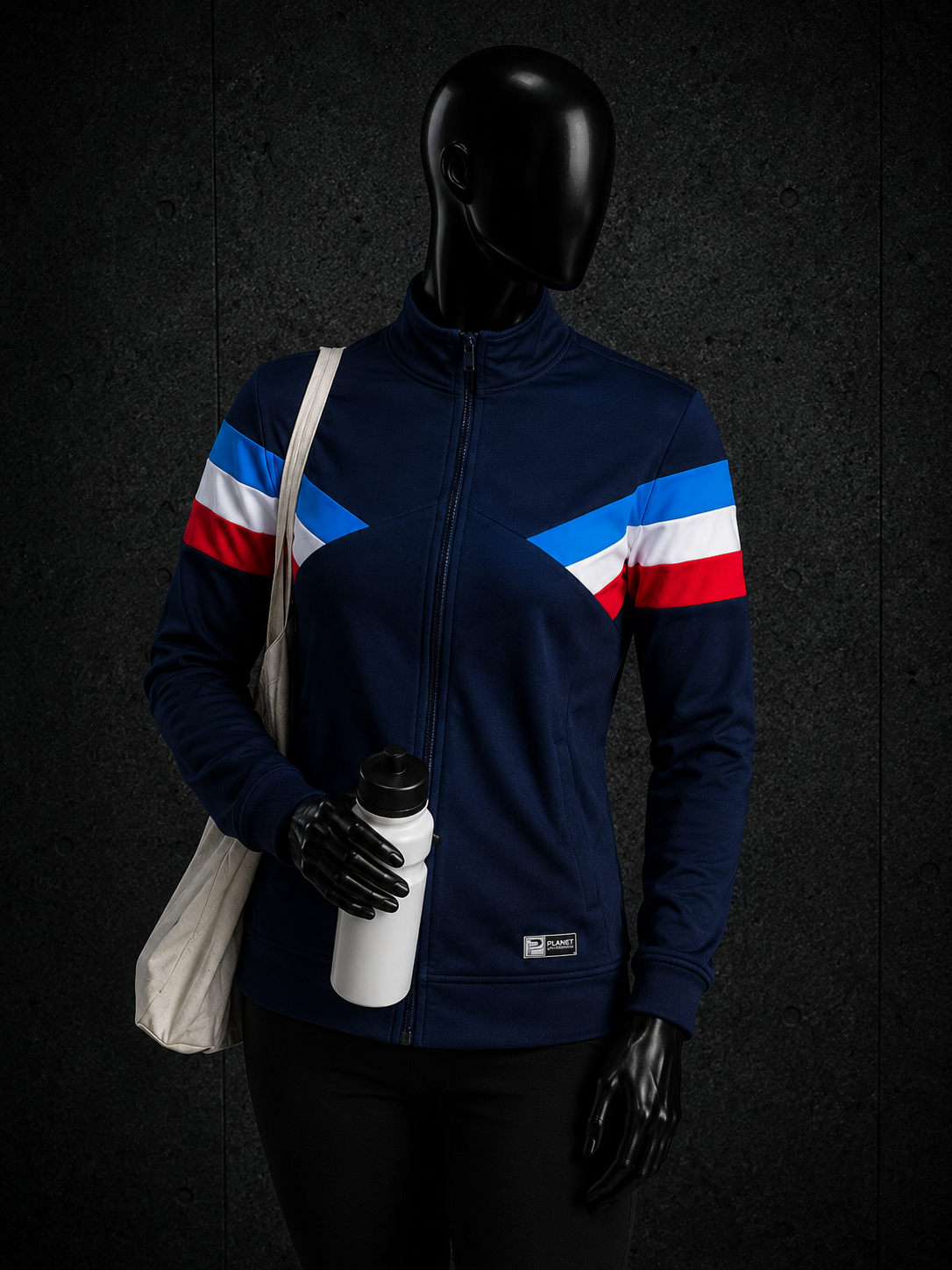 A mannequin wearing a navy blue zip-up athletic jacket with red, white, and blue diagonal stripe details on the shoulders, holding a white water bottle and a cream tote bag against a dark textured background.
