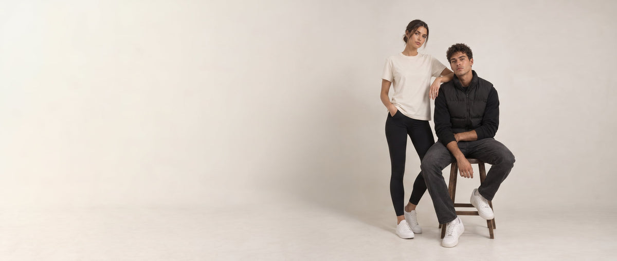 A man in a black long-sleeve shirt and dark jeans sits on a wooden stool while a woman in a white t-shirt and black leggings stands beside him, posed against a plain off-white studio background.