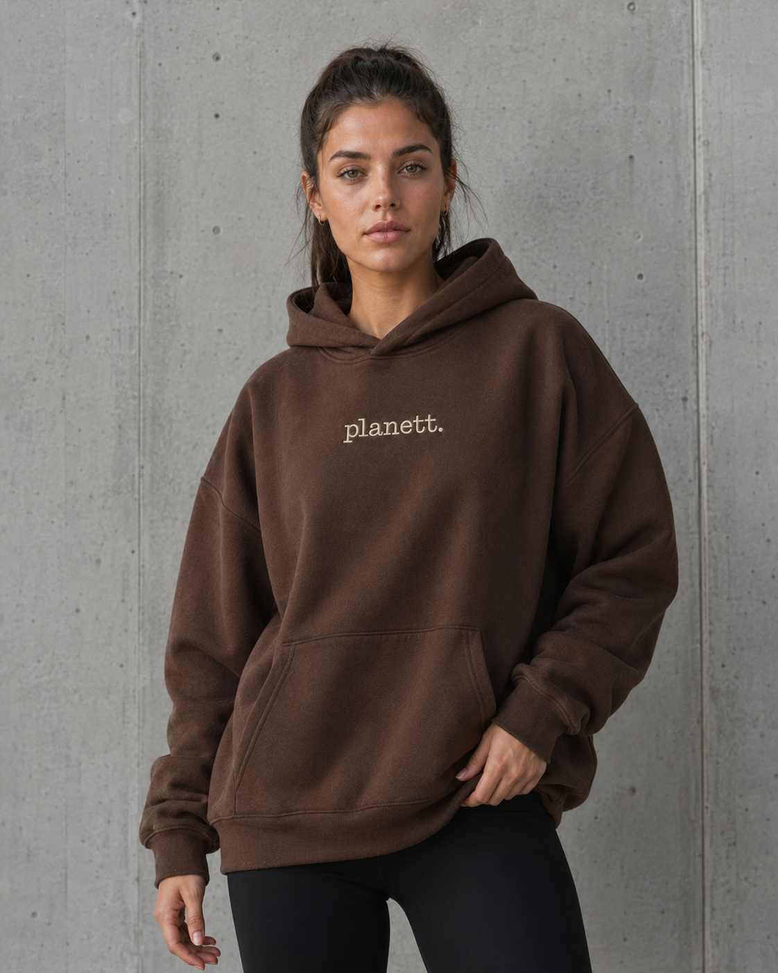 A woman wearing a brown pullover hoodie with 'planett.' embroidered in white on the chest, standing against a gray concrete wall.