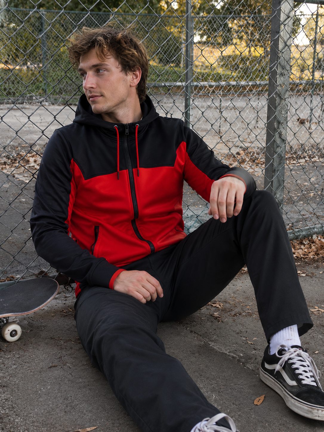 Person wearing a black and red jacket sitting on a concrete surface with a skateboard next to them.