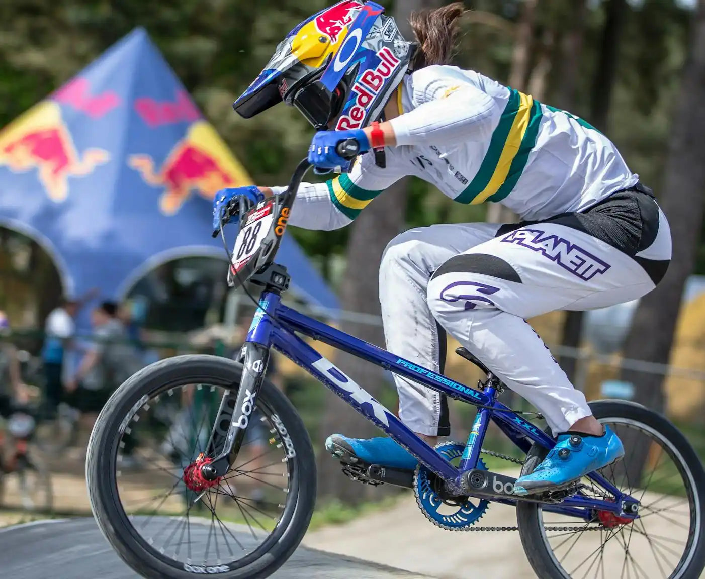 A female BMX rider in a white, green, and yellow racing uniform on a blue racing bicycle.