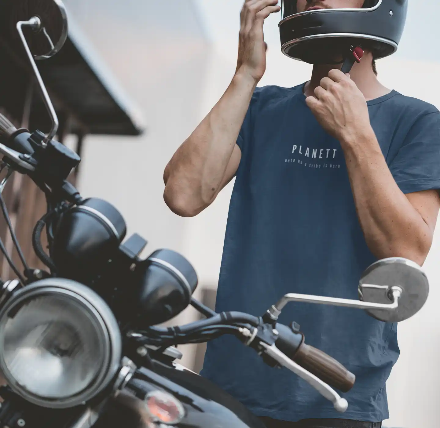 A person wearing a dark blue t-shirt and a black motorcycle helmet, adjusting the helmet’s chin strap, standing next to a vintage-style motorcycle.