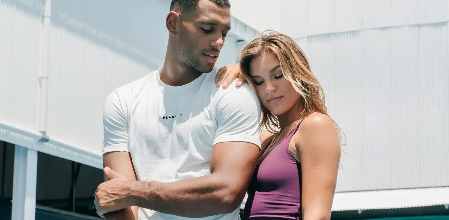 Two athletic people, a man in a white t-shirt and a woman in a purple tank top, posed intimately.