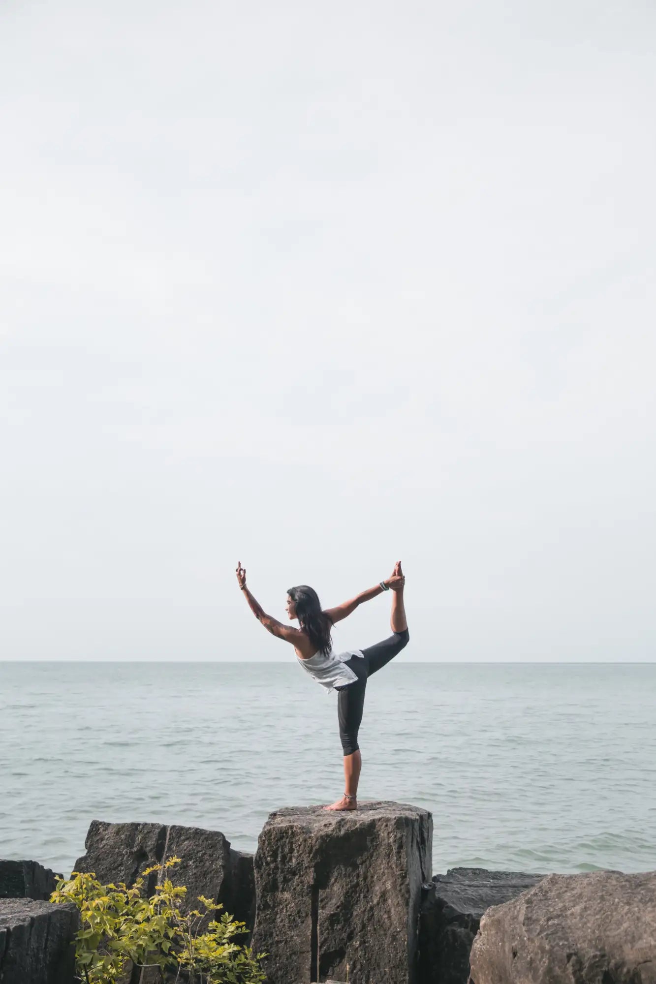 A woman in a white and gray tank top with black leggings stands barefoot on a dark, weathered rock formation by the ocean.