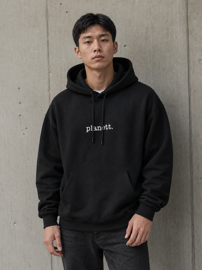 A man wearing a black pullover hoodie with 'planott.' printed in white gothic-style text on the chest, standing against a concrete wall.