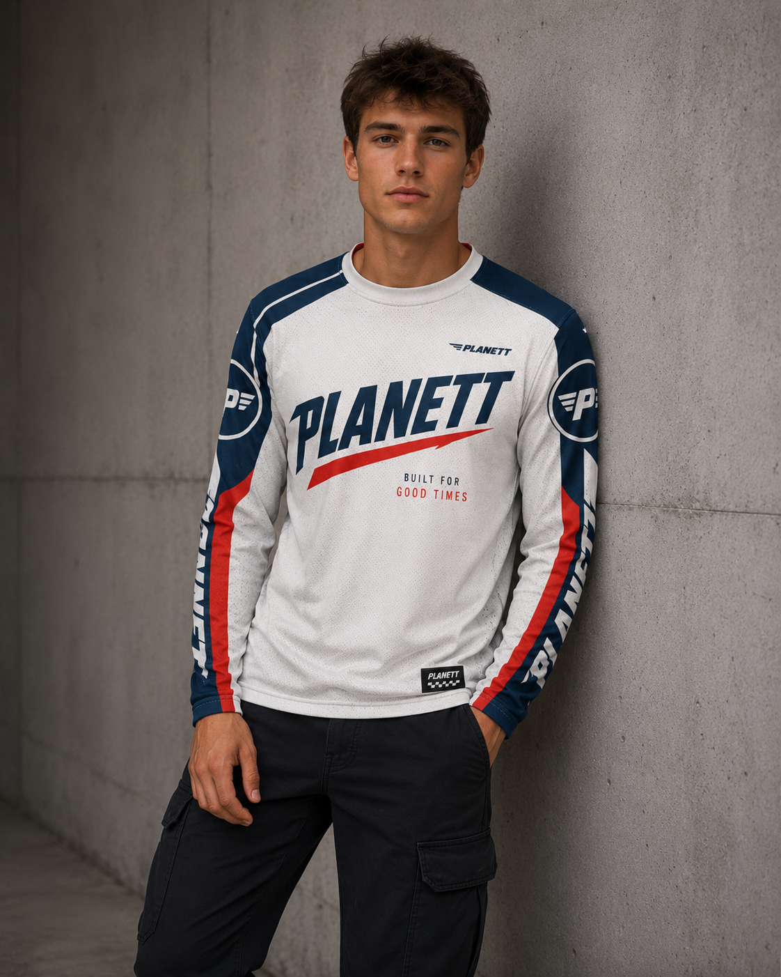 A man wearing a white long-sleeve Planett racing jersey with navy and red accents, standing against a textured gray concrete wall.