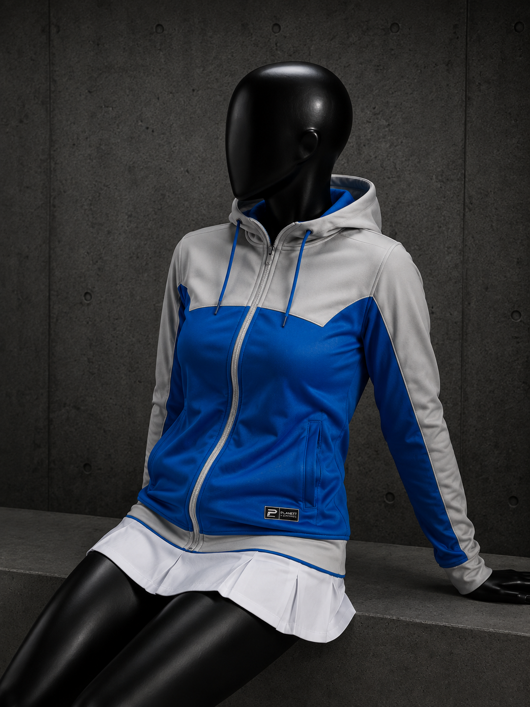 A PUMA athletic zip-up hoodie in blue and white, styled on a black mannequin with a white tennis skirt, set against a dark gray concrete wall.