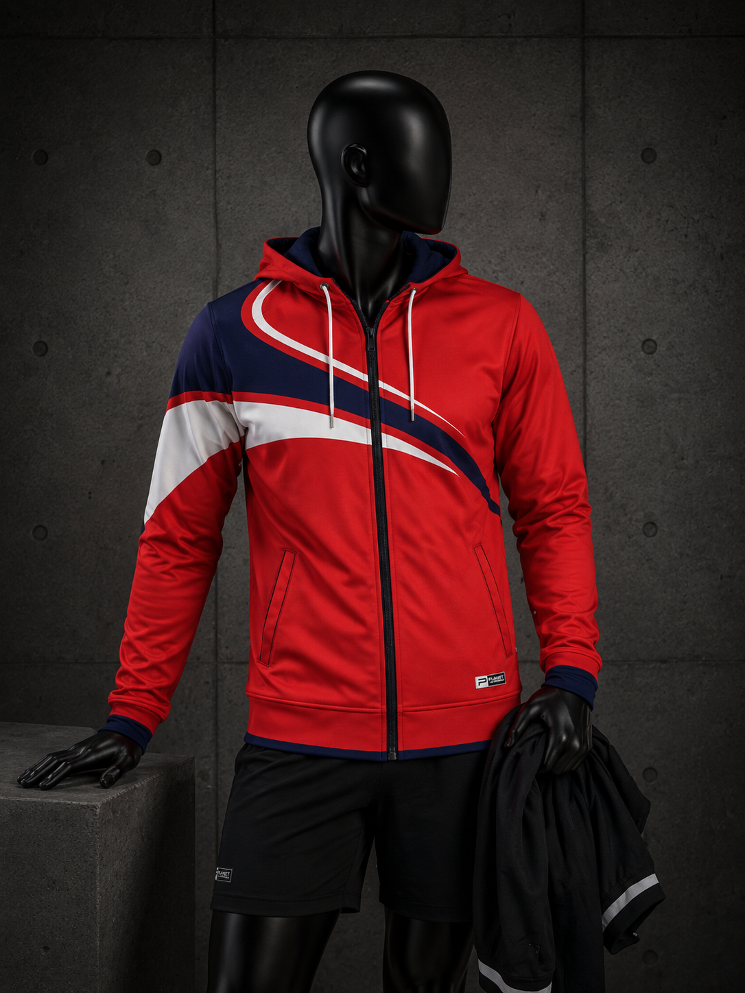 A red, navy, and white zip-up hooded sweatshirt displayed on a black mannequin against a dark gray textured wall. The hoodie features diagonal color-blocked panels in red, white, and navy, with white drawstrings and a black front zipper.