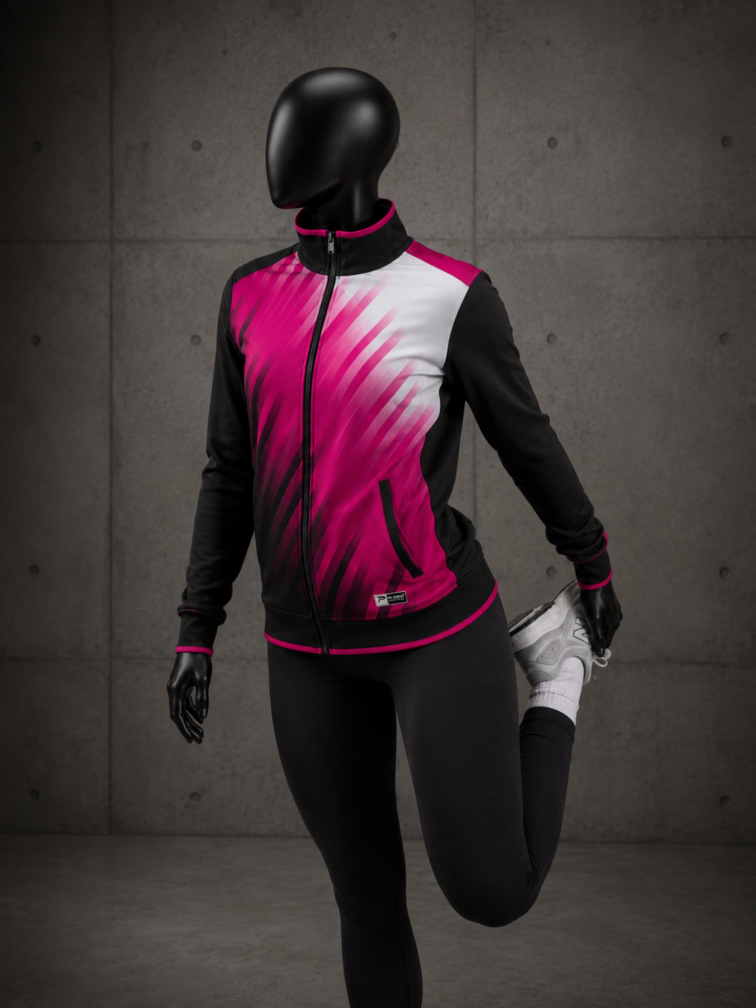 A mannequin wearing a black and bright pink athletic zip-up jacket with a white gradient panel, paired with black leggings and white gloves, standing against a gray concrete wall.