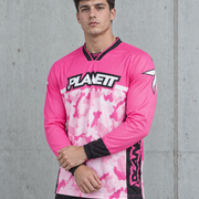 A man wearing a pink and white camouflage long-sleeve athletic jersey with black trim and the brand name PLANETT across the chest, standing against a gray concrete wall.