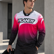 A man wearing a long-sleeve Planet brand motocross jersey with a white, pink, and black gradient design, standing outdoors in front of concrete pillars and holding a black helmet.