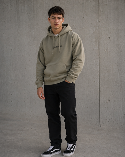 A man wearing a sage green hoodie with 'place to be' printed in black script on the chest, paired with black pants and black-and-white sneakers, standing against a concrete wall.