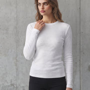 A woman modeling a white long-sleeve crewneck knit top with black athletic shorts, standing against a textured gray concrete wall.
