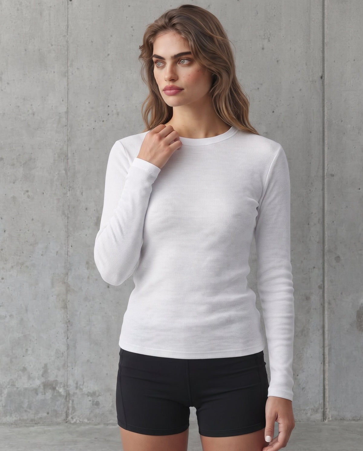 A woman modeling a white long-sleeve crewneck knit top with black athletic shorts, standing against a textured gray concrete wall.