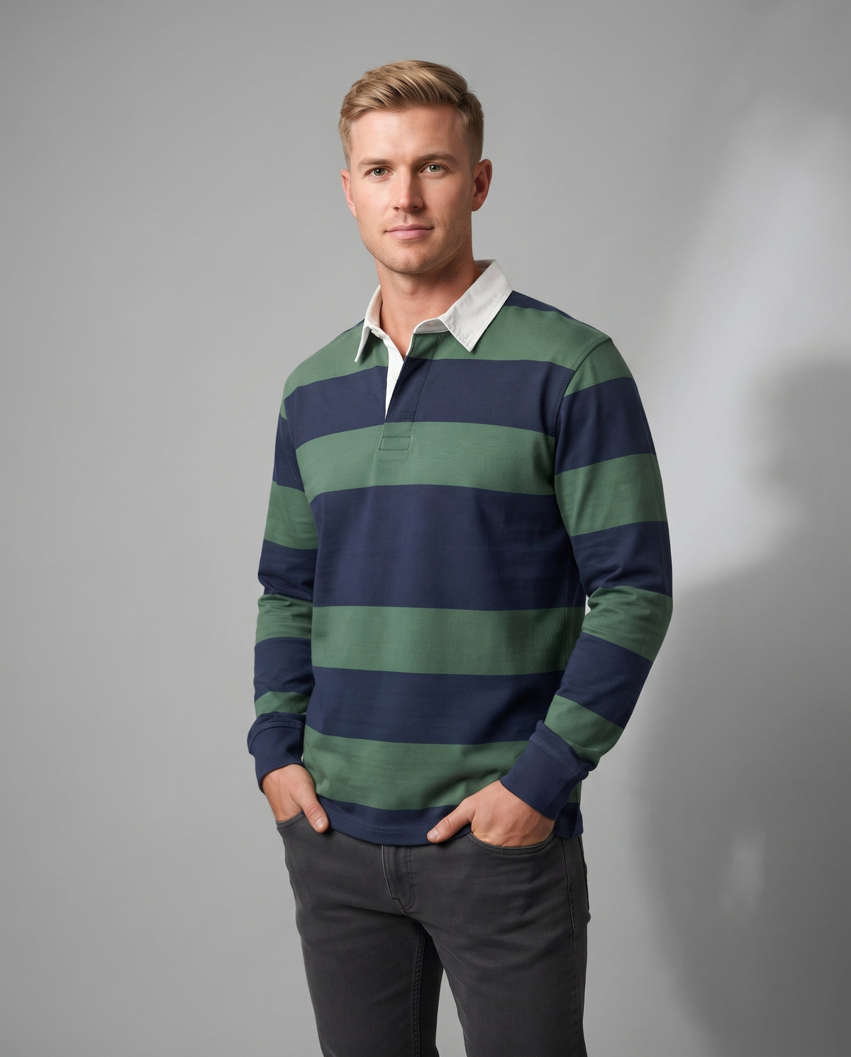 A man wearing a long-sleeve rugby shirt with horizontal navy blue and sage green stripes, paired with a white collar and dark gray pants, standing against a neutral gray studio background.