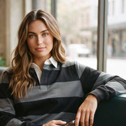 A woman sits in a teal booth at a cafe, wearing a black and gray striped long-sleeve rugby shirt over a white collared shirt. A small table in the foreground holds a cup and a notebook.