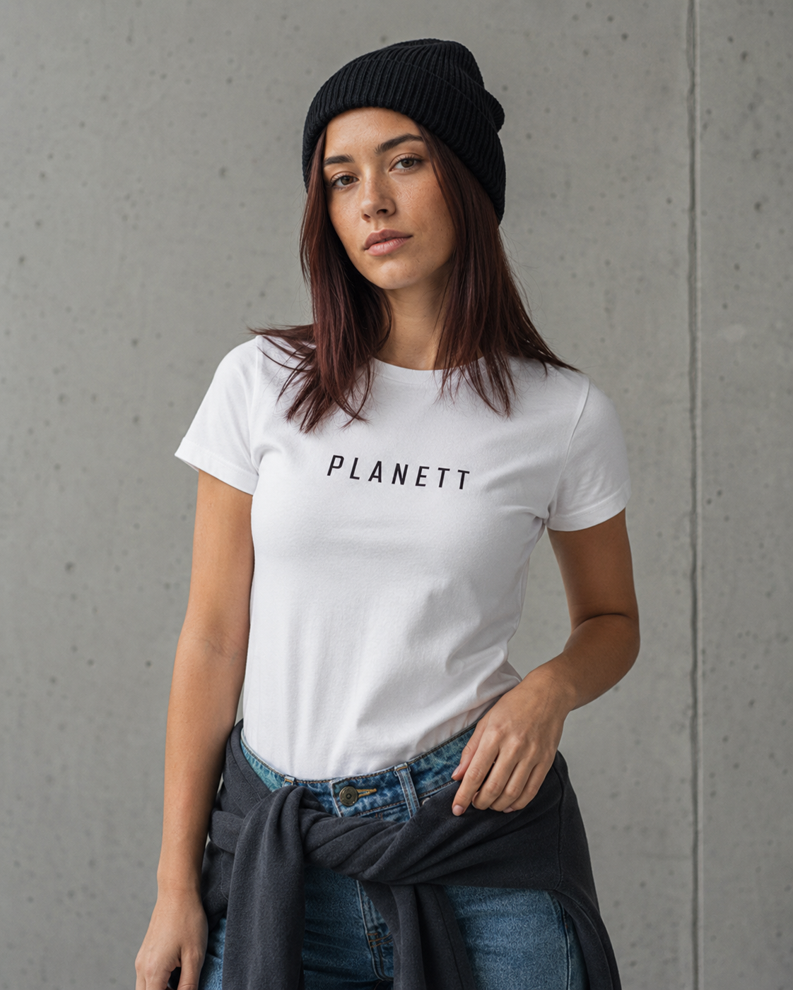 A woman wearing a white crewneck t-shirt with the brand name 'PLANETT' printed in black across the chest, styled with a black beanie, blue jeans, and a dark gray sweater tied around her waist, set against a concrete wall background.