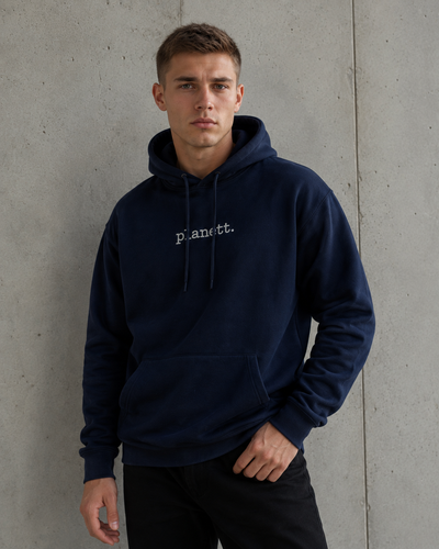 A man wearing a navy blue pullover hoodie with 'Planett.' embroidered in white on the chest, standing against a textured concrete wall.