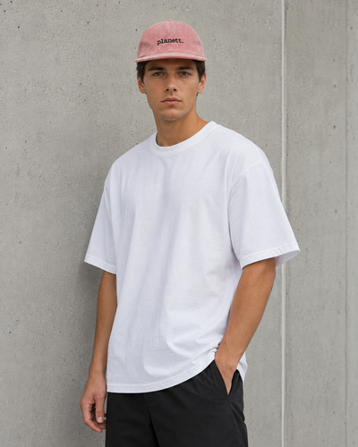 A man wearing a plain white short-sleeve crewneck t-shirt, black pants, and a pink baseball cap with the brand name 'plagent.' printed on it, standing against a textured gray concrete wall.