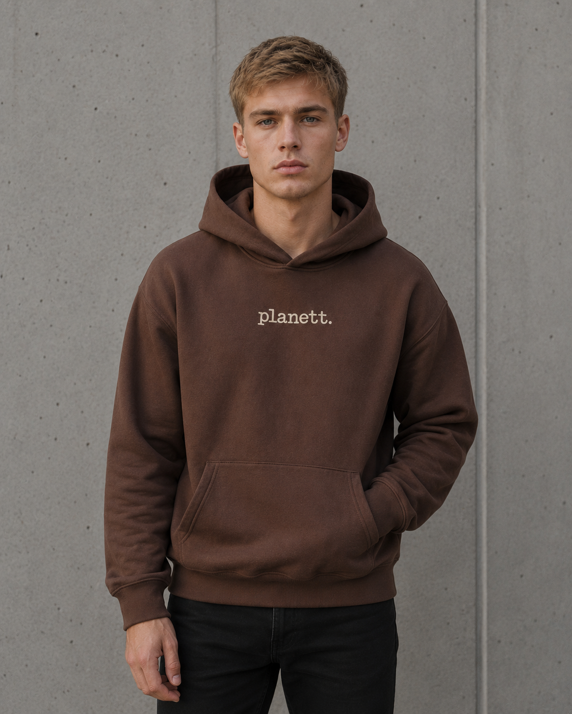 A man wearing a brown pullover hoodie with the word 'planett.' printed in white on the chest, standing against a gray concrete wall.