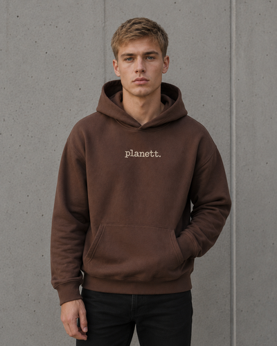 A man wearing a brown pullover hoodie with the word 'planett.' printed in white on the chest, standing against a gray concrete wall.
