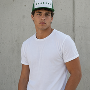 A man wearing a white crew-neck t-shirt and a white and green Planet brand trucker hat, standing against a textured concrete wall.