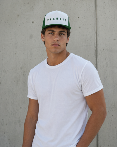 A man wearing a white crew-neck t-shirt and a white and green Planet brand trucker hat, standing against a textured concrete wall.