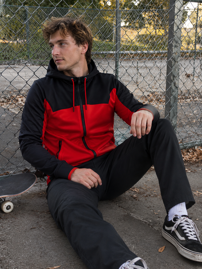 Person wearing a black and red jacket sitting on a concrete surface with a skateboard next to them.