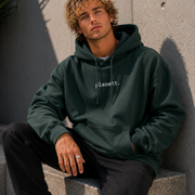 Man wearing a dark green hoodie with 'Julianett' logo, sitting against a concrete wall.