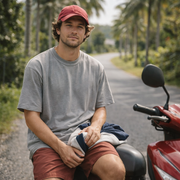 A man wearing a red baseball cap, gray t-shirt, and reddish-brown shorts sits on the seat of a red scooter.