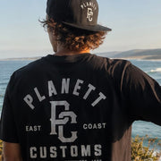 Person wearing a black 'Planett East Coast Customs' t-shirt and cap, standing by the ocean.