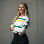 A smiling woman with long blonde hair wearing a white long-sleeved shirt with green and yellow stripes, and black pants.