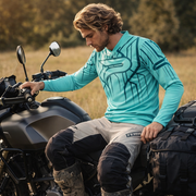 A man wearing a turquoise long-sleeved motocross jersey with black geometric patterns and the fox logo, paired with beige and black riding pants.
