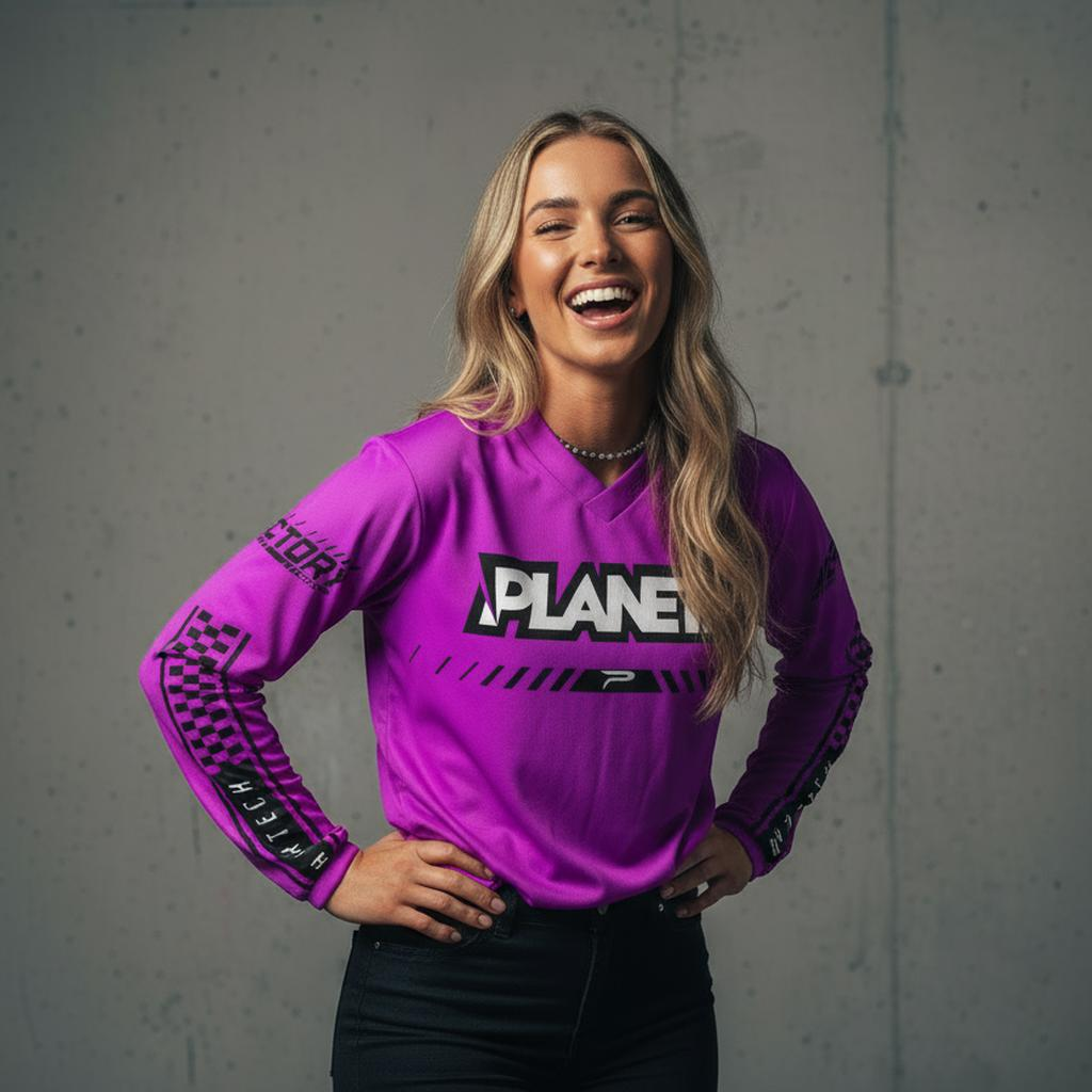 A smiling woman with long blonde hair wearing a long-sleeved purple shirt with black checkered patterns on the sleeves and black pants.