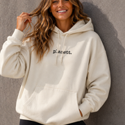 Woman wearing a beige hoodie with 'p.anett.' text against a gray wall