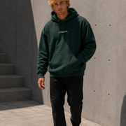 Person wearing a green hoodie with text, standing against a concrete wall.