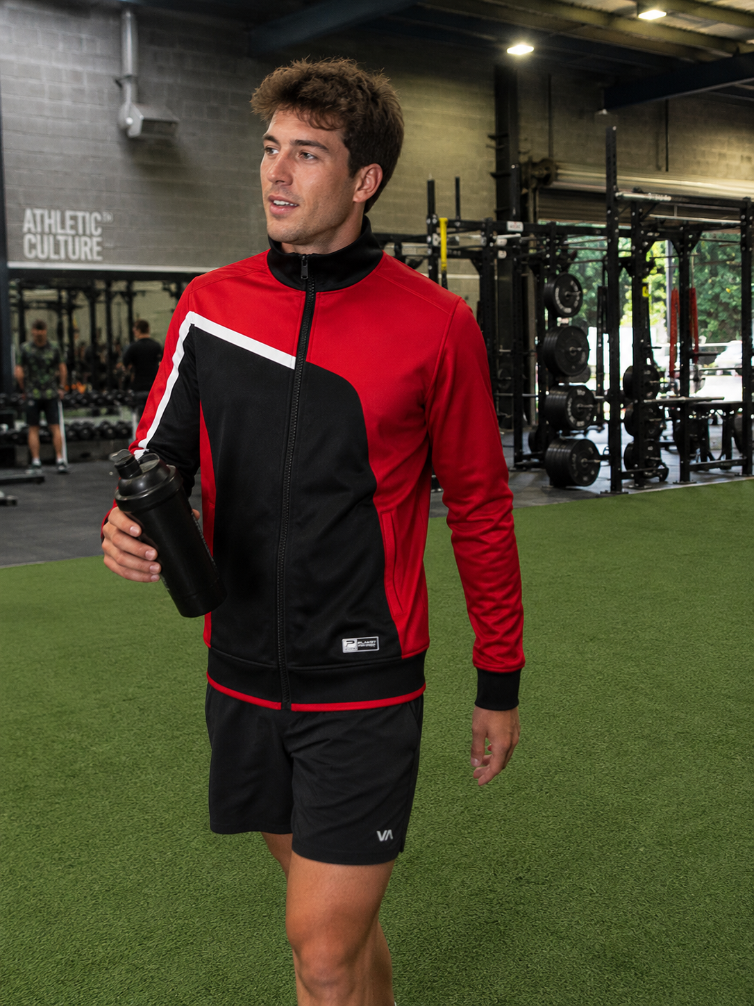Man in a red and black athletic outfit holding a water bottle in a gym.