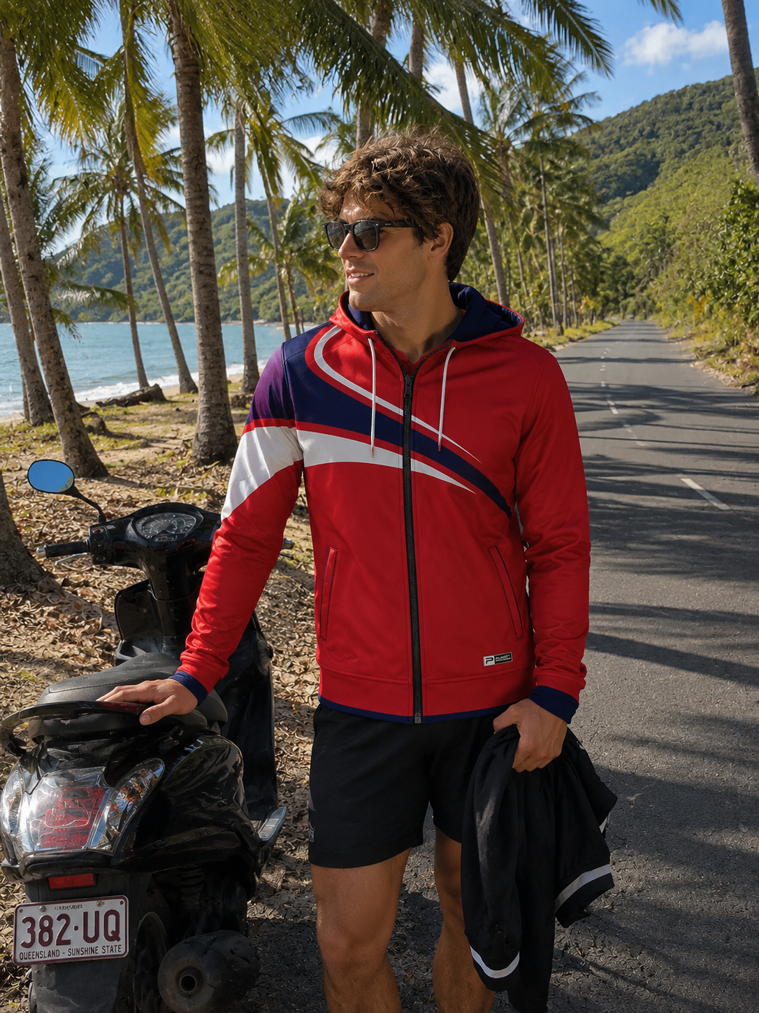 Man in a red jacket standing next to a four-wheeler on a scenic road with palm trees and mountains.