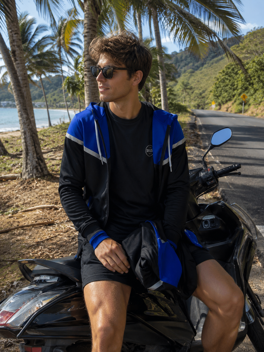 Man sitting on a motorcycle by a scenic lakeside with palm trees and mountains in the background.