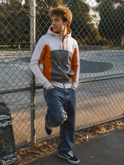 Person wearing a colorful hoodie and jeans leaning against a chain-link fence with skateboards in the background.