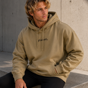 Man wearing a beige hoodie with text, sitting against a concrete wall.