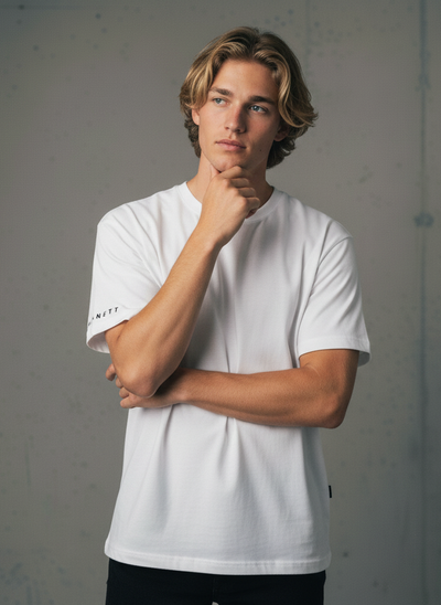 A young man with shoulder-length blonde hair wearing a white short-sleeve t-shirt and black pants.