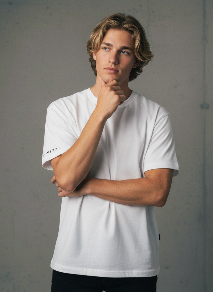 A young man with shoulder-length blonde hair wearing a white short-sleeve t-shirt and black pants.