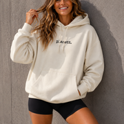 Woman wearing a white hoodie with text, standing against a gray wall.
