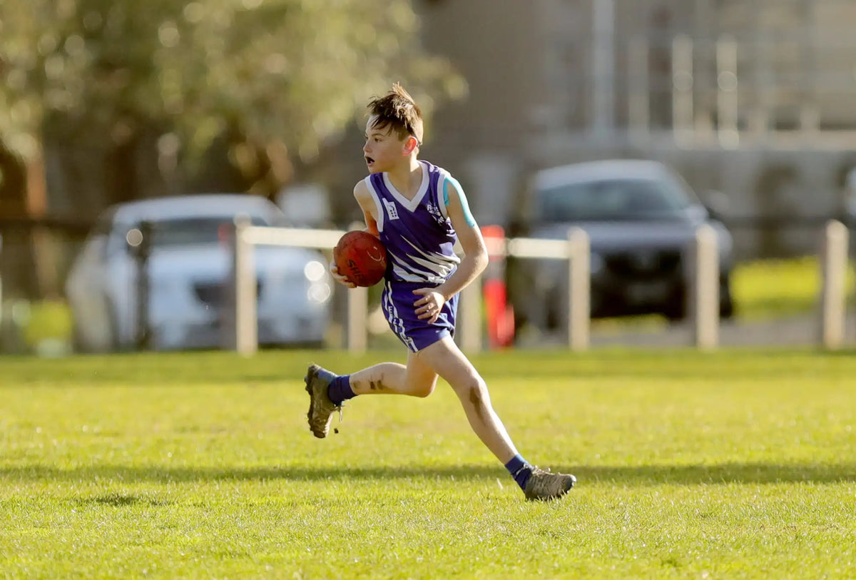 A young boy in a purple and white striped athletic jersey runs across a grassy field while holding an orange football.